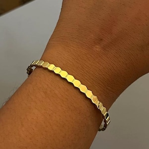May include: A gold-colored bracelet with a honeycomb design. The bracelet is made of small, connected hexagonal shapes. The jewelry is worn on a wrist, and the background is a neutral color.