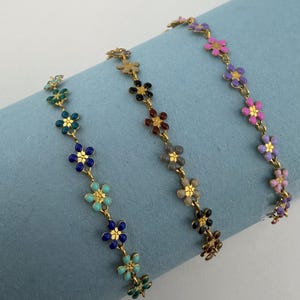 Könnte beinhalten: Drei goldfarbene Armbänder, jedes mit bunten Emaille-Blütenanhängern. Die Armbänder zeigen blaue, grüne, rosa, lila und braune Blumen. Das florale Design verleiht einen Hauch von Eleganz.