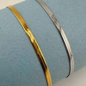 May include: Two flat, herringbone-style necklaces. One is gold-coloured, the other is silver-coloured. Both are displayed on a light blue surface. The necklaces have a smooth, reflective finish.