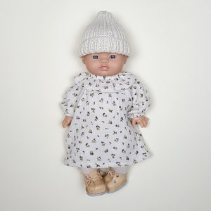 May include: A doll wearing a white knitted beanie, a white dress with a floral pattern, and brown shoes.