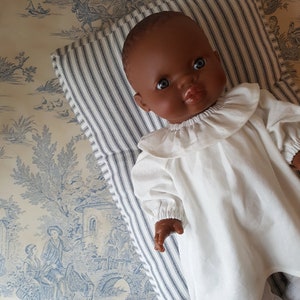 May include: A dark-skinned baby doll wearing a white ruffled dress with long sleeves. The doll is lying on a striped pillow.