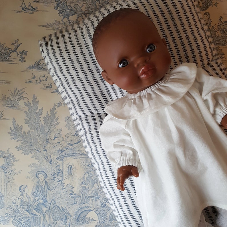 May include: A dark-skinned baby doll wearing a white ruffled dress with long sleeves. The doll is lying on a striped pillow.