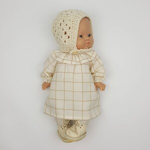 May include: A doll wearing a cream-coloured dress with a plaid pattern, a cream-coloured crocheted hat, and white shoes.