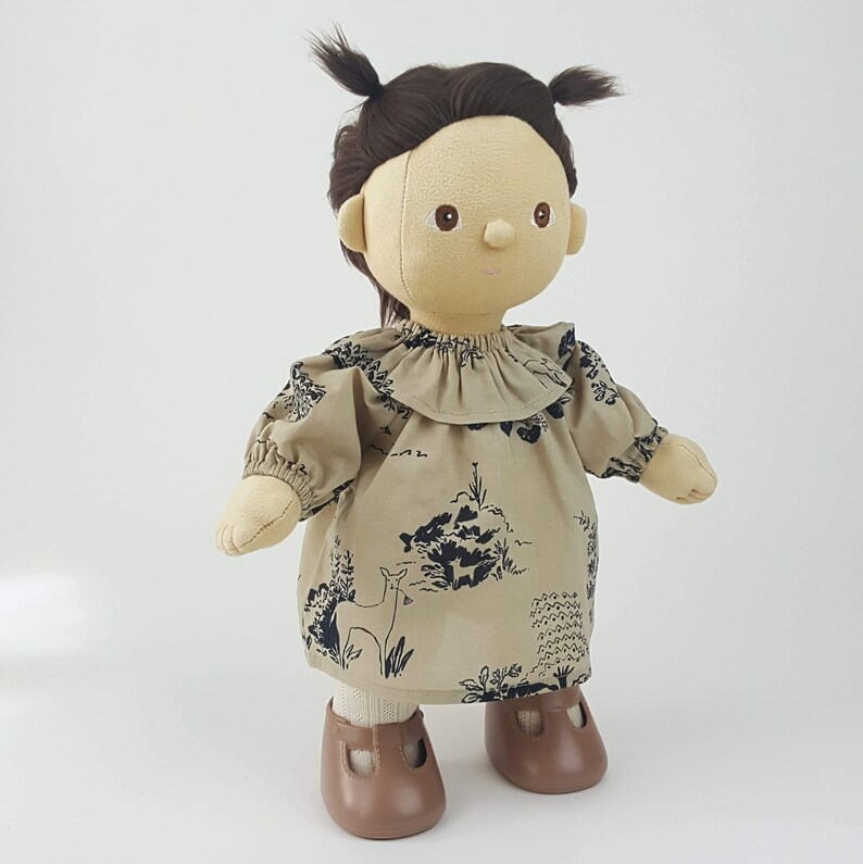 May include: A brown doll with brown hair in pigtails wearing a beige dress with a black forest scene and a black deer print. The doll is wearing brown shoes.
