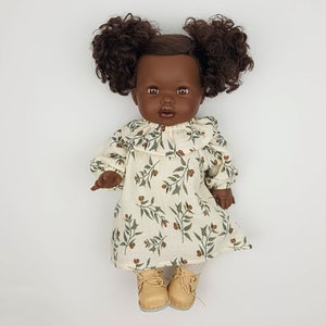 May include: A doll with brown curly hair wearing a white dress with a green and brown floral pattern and brown shoes.