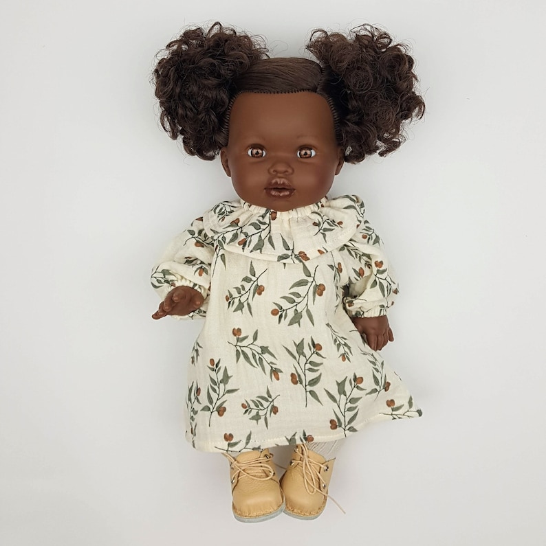 May include: A doll with brown curly hair wearing a white dress with a green and brown floral pattern and brown shoes.