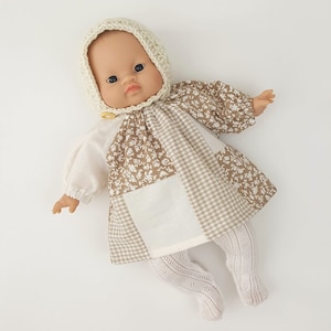 Op de afbeelding: Een pop met een crèmekleurige gehaakte muts en een patchworkjurk met bruine en witte bloemen- en ruitpatronen. De pop draagt witte kousen met een kantpatroon.