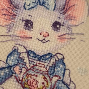May include: A cross-stitch embroidery on white fabric featuring a light purple mouse with large dark eyes, pink cheeks, and a blue bow on its head. The mouse wears a blue dress with a white apron and holds a small teapot with a floral pattern.