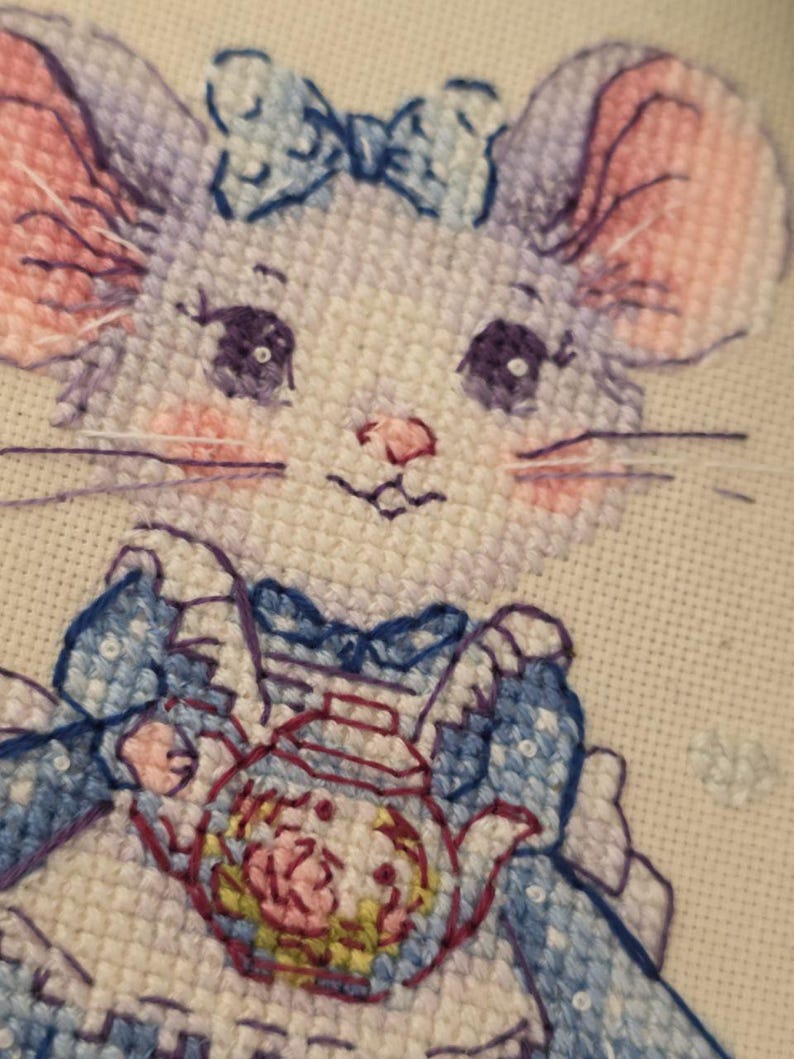 May include: A cross-stitch embroidery on white fabric featuring a light purple mouse with large dark eyes, pink cheeks, and a blue bow on its head. The mouse wears a blue dress with a white apron and holds a small teapot with a floral pattern.