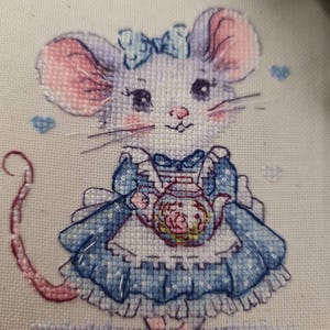 May include: A cross-stitch embroidery on off-white fabric depicting a mouse with large pink-tinted ears, wearing a blue dress with a white apron and a blue bow. The mouse holds a small teapot with a floral design, surrounded by tiny blue hearts.