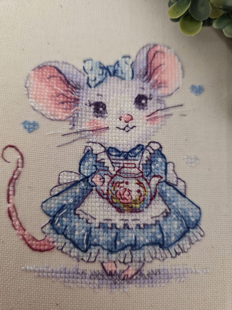 May include: A cross-stitch embroidery on off-white fabric depicting a mouse with large pink-tinted ears, wearing a blue dress with a white apron and a blue bow. The mouse holds a small teapot with a floral design, surrounded by tiny blue hearts.