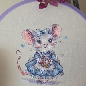 May include: A cross-stitch embroidery in a purple circular hoop featuring a mouse with pink ears and cheeks, wearing a blue dress with a white apron and a blue bow on its head. The mouse holds a small teapot, surrounded by tiny blue hearts.