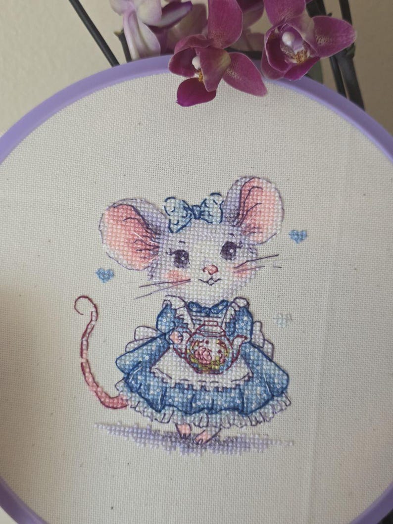 May include: A cross-stitch embroidery in a purple circular hoop featuring a mouse with pink ears and cheeks, wearing a blue dress with a white apron and a blue bow on its head. The mouse holds a small teapot, surrounded by tiny blue hearts.