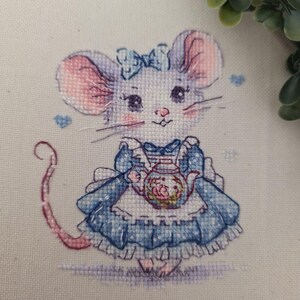 May include: A cross-stitch embroidery on cream fabric featuring a mouse wearing a blue dress with a white apron and a blue bow on its head. The mouse holds a small teapot with floral details. Small blue hearts are stitched around the mouse.