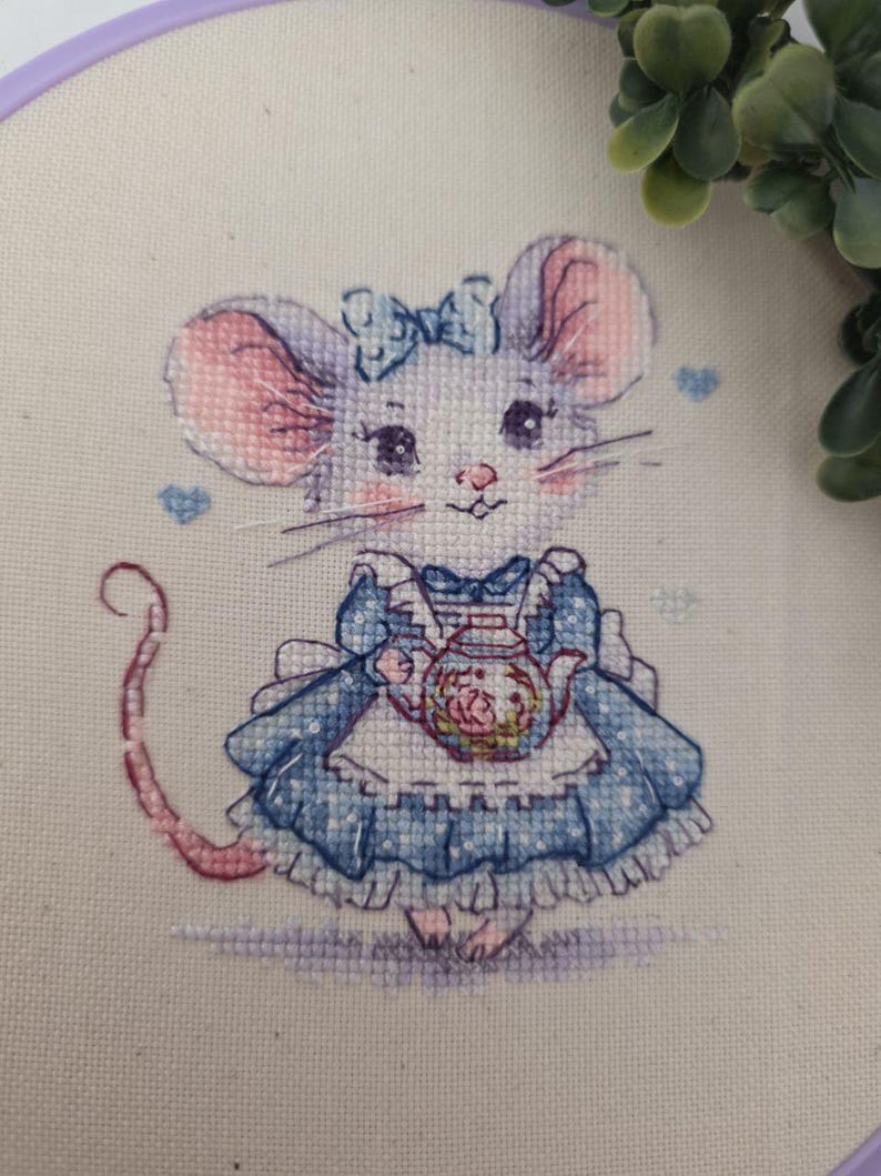 May include: A cross-stitch embroidery on cream fabric featuring a mouse wearing a blue dress with a white apron and a blue bow on its head. The mouse holds a small teapot with floral details. Small blue hearts are stitched around the mouse.