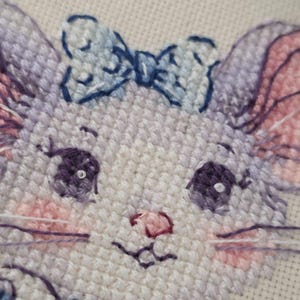 May include: A cross-stitch embroidery of a cat face featuring light purple fur, large dark purple eyes with white highlights, pink cheeks, and a small pink nose. The cat wears a light blue bow on its head and a blue collar, stitched on white fabric.
