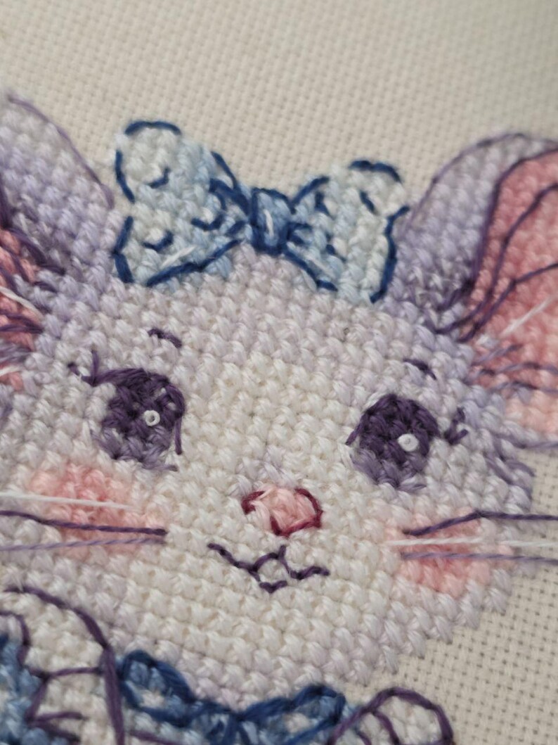 May include: A cross-stitch embroidery of a cat face featuring light purple fur, large dark purple eyes with white highlights, pink cheeks, and a small pink nose. The cat wears a light blue bow on its head and a blue collar, stitched on white fabric.