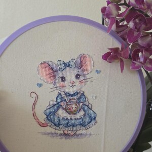 May include: A cross-stitch embroidery piece in a round purple hoop featuring a mouse wearing a blue dress with a white apron and a blue bow on its head. The mouse is holding a small teapot. The design is stitched on off-white fabric.