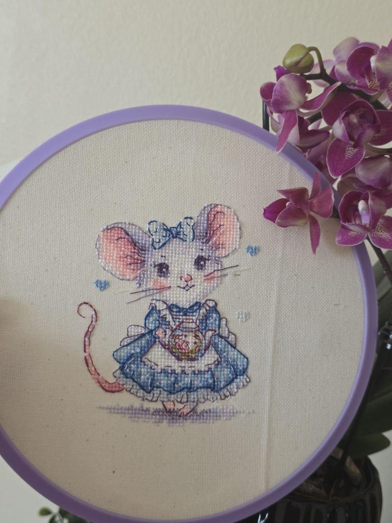 May include: A cross-stitch embroidery piece in a round purple hoop featuring a mouse wearing a blue dress with a white apron and a blue bow on its head. The mouse is holding a small teapot. The design is stitched on off-white fabric.
