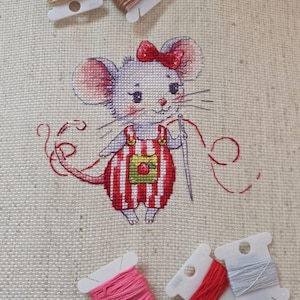 Peut inclure: Une broderie au point de croix sur tissu beige représentant une souris grise portant une salopette rayée rouge et blanche avec un écusson pomme verte et un nœud rouge. La souris tient une aiguille avec du fil rouge, entourée de cinq bobines.
