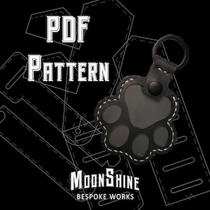 Può includere: Portachiavi a forma di zampa di animale in pelle nera con cuciture bianche e anello in metallo. L'immagine include il testo "PDF PATTERN" e "MOONSHINE BESPOKE WORKS" su sfondo nero con un motivo.