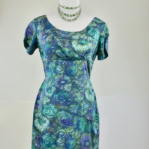 Puede incluir: Vestido de estilo vintage con estampado floral en tonos turquesa, azul y verde. El vestido presenta un escote redondo, mangas cortas y una silueta ajustada. También se muestra un collar de cuentas verdes a juego.