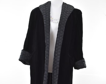 May include: A long, black velvet coat with a quilted satin trim along the collar, lapels, and cuffs. The coat is displayed on a wooden dress form against a white background. The coat has a classic, elegant design.