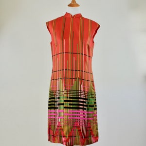 May include: A sleeveless, knee-length dress in a vibrant orange hue, adorned with vertical stripes in various colors. The dress features a mandarin collar and is displayed on a wooden dress form. The bottom of the dress has a geometric pattern.
