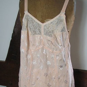 May include: A vintage pink lace and satin corset with floral embroidery and adjustable straps. The corset has six garter straps and is displayed on a mannequin.