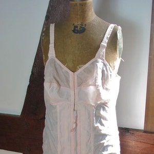 May include: A vintage pink nylon corset with a zipper closure and adjustable straps. The corset is displayed on a wire dress form.