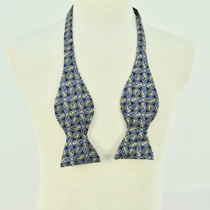 May include: A navy blue bow tie with a repeating pattern of yellow, green, and light blue circles and curved shapes. The bow tie is displayed on a white mannequin, showcasing its design and shape.