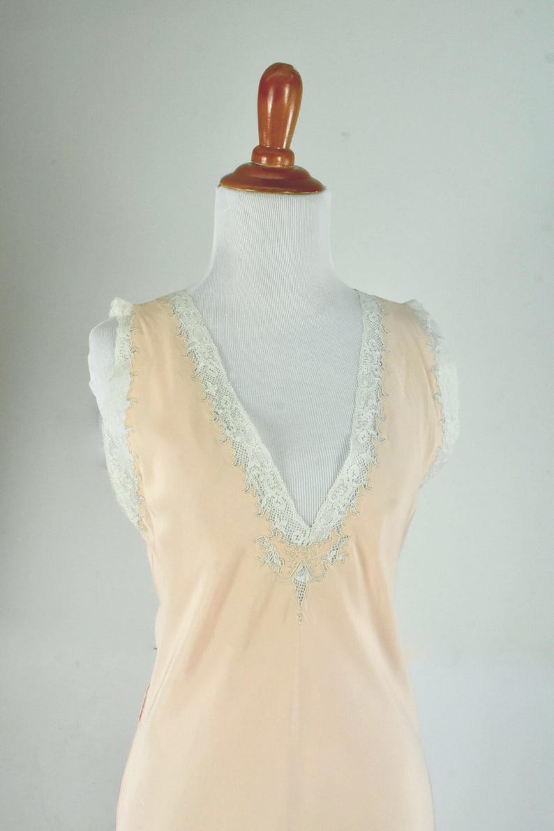 May include: A peach-colored, sleeveless slip dress with a V-neckline. The neckline and armholes are trimmed with white lace. The dress is displayed on a mannequin.