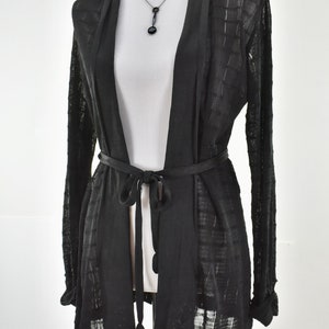 May include: A black open-front cardigan with long sleeves and a matching belt tied at the waist. The cardigan is displayed on a white mannequin. A black necklace with a pendant is also visible.
