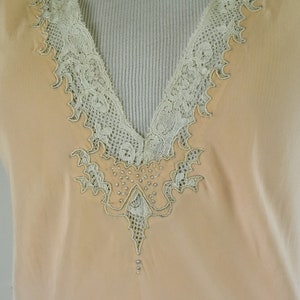 May include: A peach-colored sleeveless top with a V-neckline. The neckline and edges are trimmed with delicate, off-white lace. The lace design extends down the front, embellished with small, shiny beads.