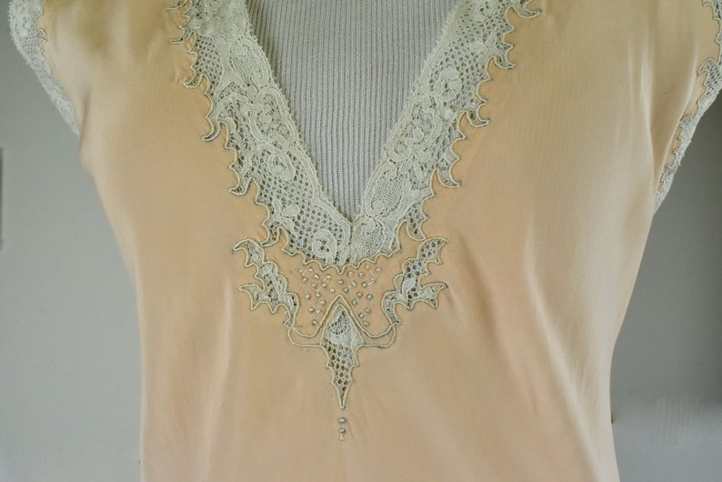May include: A peach-colored sleeveless top with a V-neckline. The neckline and edges are trimmed with delicate, off-white lace. The lace design extends down the front, embellished with small, shiny beads.
