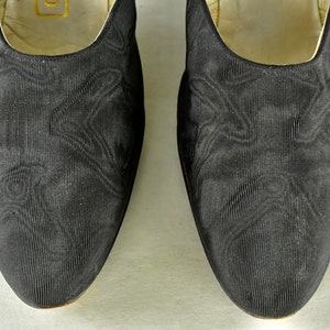 May include: A pair of black satin slippers with a subtle textured pattern. The slippers have a pointed toe and a low heel.
