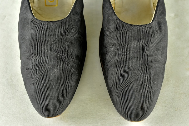 May include: A pair of black satin slippers with a subtle textured pattern. The slippers have a pointed toe and a low heel.