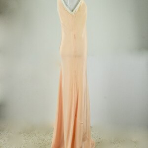 May include: A peach-colored, floor-length slip dress on a mannequin. The dress features a V-neckline trimmed with white lace and a flowing skirt. The fabric appears to be a lightweight material, ideal for formal occasions.