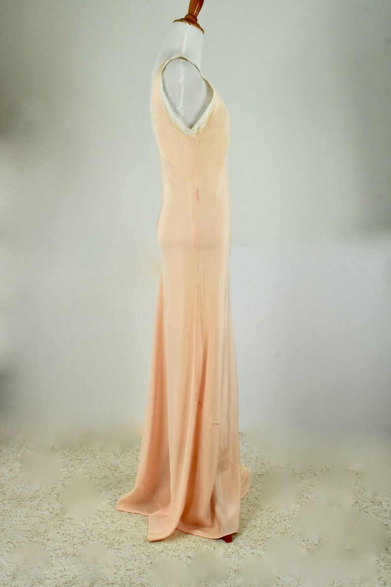 May include: A peach-colored, floor-length slip dress on a mannequin. The dress features a V-neckline trimmed with white lace and a flowing skirt. The fabric appears to be a lightweight material, ideal for formal occasions.