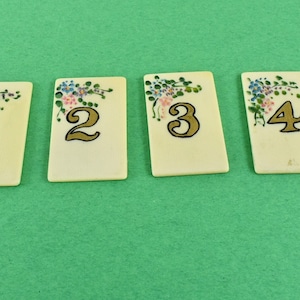 May include: Four ivory-colored table number cards, numbered 1 through 4 in gold, with floral corner designs in pink, blue, and green. The cards are arranged on a solid green background.
