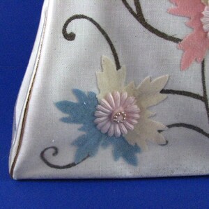 May include: A white fabric handbag with a triangular shape and a floral design. The flowers are made of fabric and are in shades of pink, blue, and white.