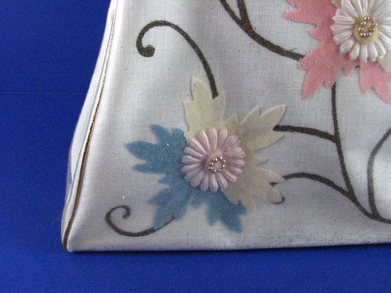 May include: A white fabric handbag with a triangular shape and a floral design. The flowers are made of fabric and are in shades of pink, blue, and white.