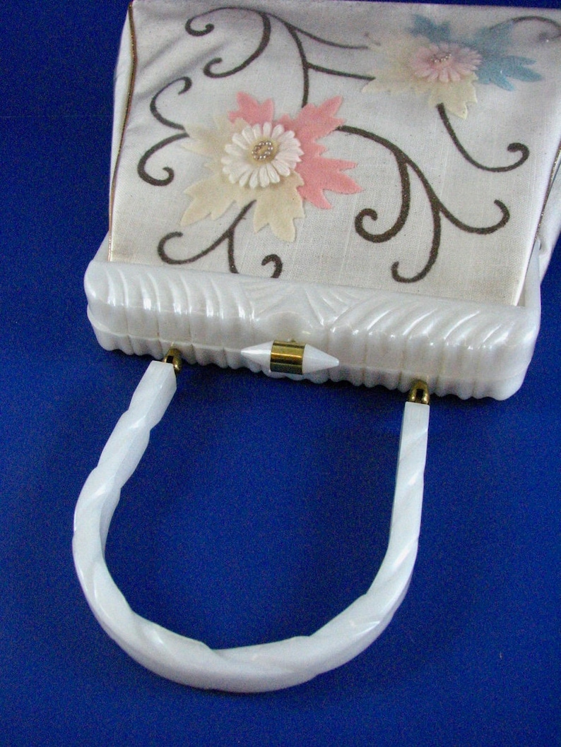 May include: White vintage handbag with a white plastic handle and a floral design on the front. The flowers are pink, white, and blue.