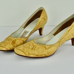May include: A pair of light tan leather pumps with floral embossed detailing. The shoes have a pointed toe, a low heel, and a white insole. The label inside the shoe reads "Rene Mancini".