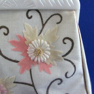 May include: A white fabric handbag with a floral design. The flowers are made of felt and are in shades of pink, white, and cream. The handbag has a white frame and a gold trim.