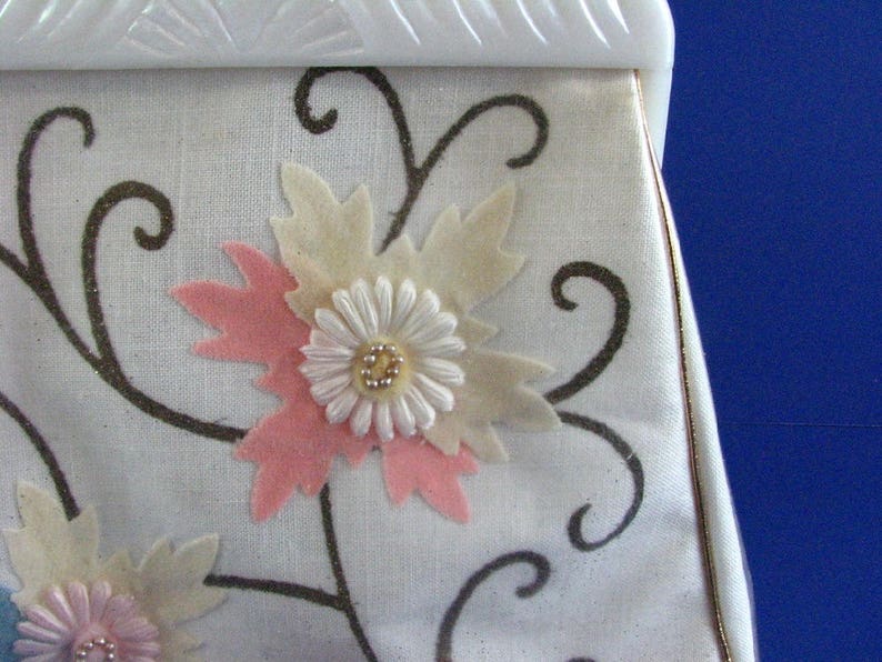 May include: A white fabric handbag with a floral design. The flowers are made of felt and are in shades of pink, white, and cream. The handbag has a white frame and a gold trim.