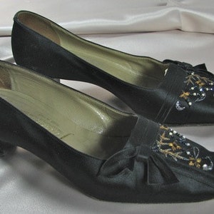 Puede incluir: Un par de zapatos de satén negro con tacón bajo. Los zapatos están decorados con un diseño floral y cuentas.