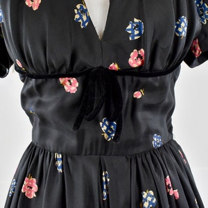 Op de afbeelding: Een zwarte jurk met een bloemenprint en een strik van fluweel in de taille.