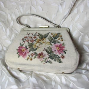 Puede incluir: Bolso vintage color crema con diseño floral de punto de aguja. El bolso presenta un asa superior, un cierre dorado y un patrón bordado detallado de flores rosas, amarillas y verdes. El bolso está sobre un fondo blanco.