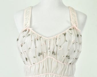 May include: A vintage-style, light pink nightgown on a mannequin. The sheer bodice features a floral pattern and is trimmed with pink piping. The straps and waistline are also accented with pink. The skirt is a flowing, opaque white.